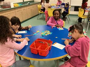 Four students at a round table use pieces of construction paper to create mosaics in the style of Alma Thomas.