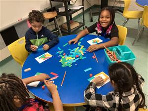 Four students at a round table use pieces of construction paper to create mosaics in the style of Alma Thomas.