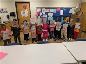 Picture shows a group of pre-k students lined up, holding up their colored pictures of paper masks they created.