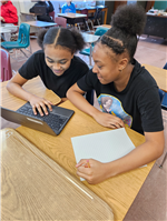 Two female students at a desk, using a computer to research a STEM pioneer.