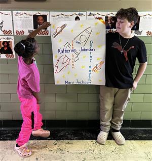 A female and male student holding up a graphic showing the interconnectedness of several female scientists to John Glenn.