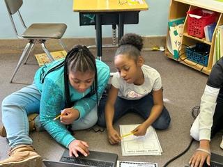 Two female Perry Elementary students sit on the floor, huddled over a laptop, working together on a research project.