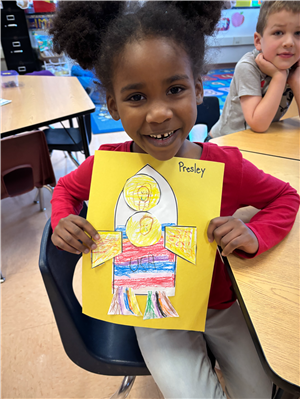 A little girl shows off construction paper artwork featuring a rocketship.