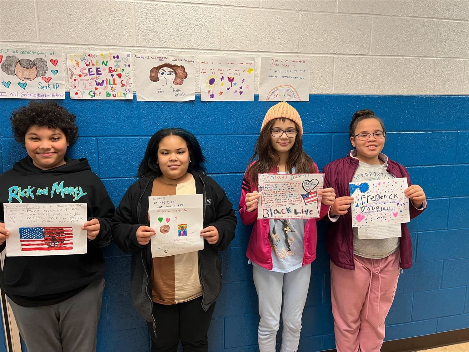 Four students lined up against a hallway wall holding up decorated paper reports on famous Black politicians.