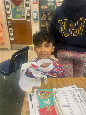 A male student sitting at a desk displaying a paper project meant to resemble an invention made by a Black American in STEM.