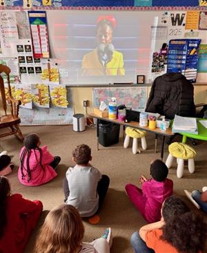 Students sitting on a classroom floor watching a screen showing poet Amanda Gorman reading at the presidential inauguration.