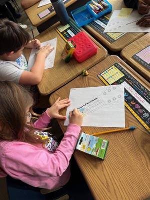 Photo shows two students at a desk, coloring a picture of a part of a guitar.